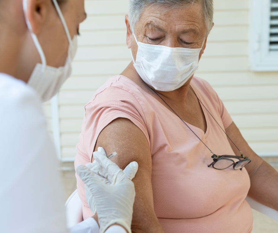 Image of a nurse with short black hair giving a homebound senior with white hair a vaccine. Both are masked.