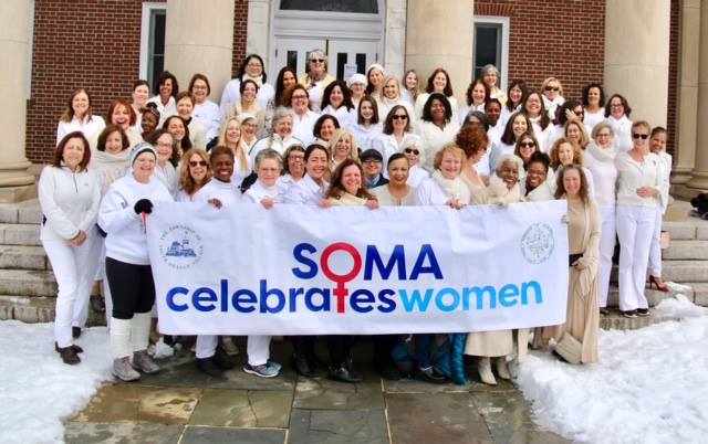 Photo of many women from the SOMA community, all wearing white, holding a 