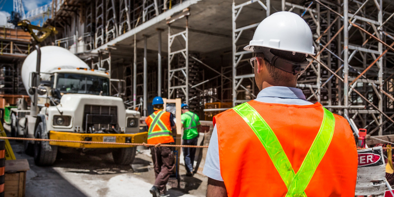 Image of a construction worker in an orange vest and white hard hat. There is a construction truck, a building, and additional workers visible in the background.