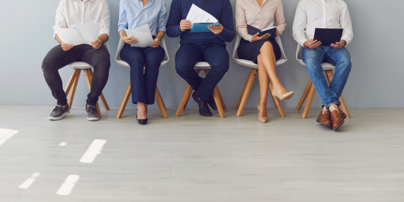 Image of five job seekers waiting in chairs, cropped from the waist down. The five individuals are of different races and are wearing different business casual outfits in shades of blue and beige.