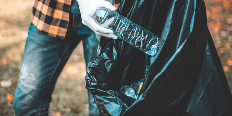 Image of an individual in an orange plaid shirt, jeans, and plastic gloves picking up a piece of litter (clear plastic bottle) from the grass and placing it into a trash bag.