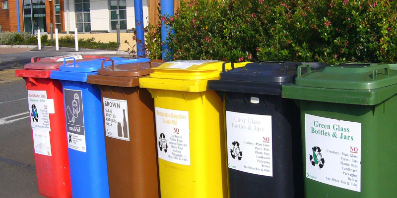 Image of different color-coded recycling bins for various types of waste. There is a red bin, a blue bin, a brown bin, a yellow bin, a black bin, and a green bin in a row on top of asphalt. There is a brick building and hedges in the background.