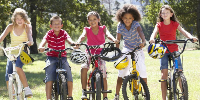 Image of five children of different races and genders on colorful bikes. In the background there is grass and trees.