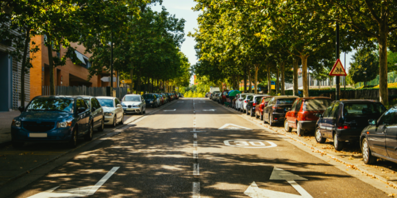 Image of a two-way, tree-lined street. There are cars parked on either side of the road.