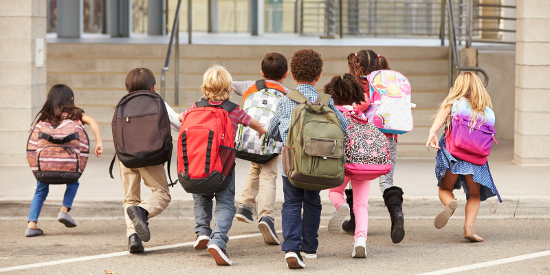 Image of a diverse group of children in colorful backpacks walking into the front of a school.